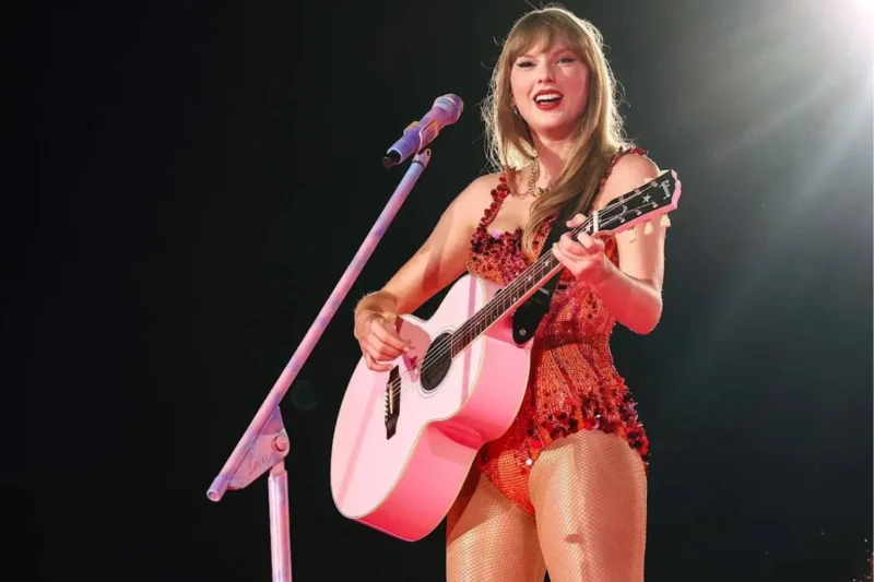 Taylor Swift performing live on stage during The Eras Tour, wearing a sparkly pink and red bodysuit and playing a pastel pink acoustic guitar into a matching pink microphone stand.