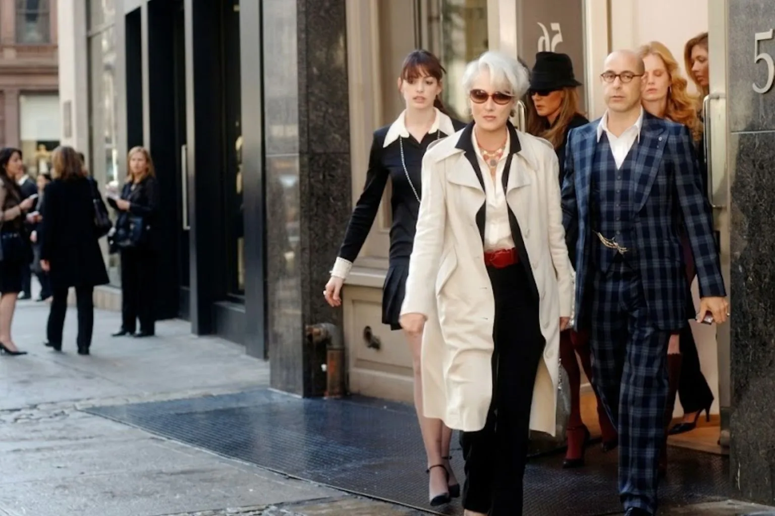Meryl Streep as Miranda Priestly in a white trench coat, walking down a New York street flanked by Anne Hathaway and Stanley Tucci in a scene from The Devil Wears Prada.