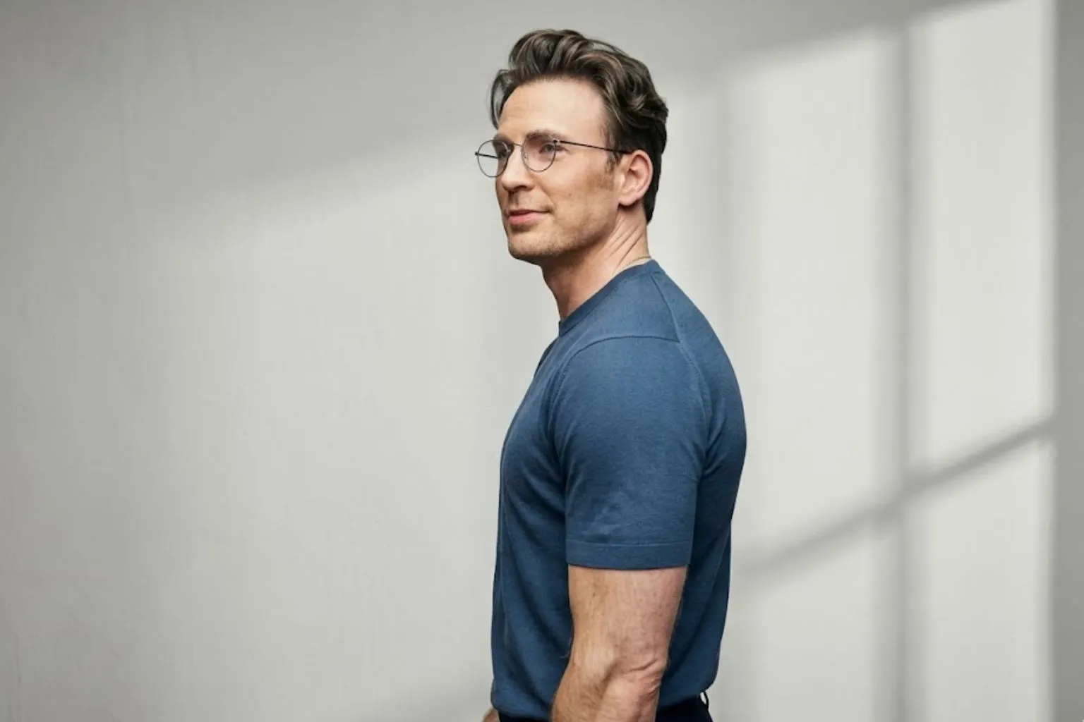 Chris Evans wearing a blue t-shirt and thin-frame glasses against a minimalist grey background.