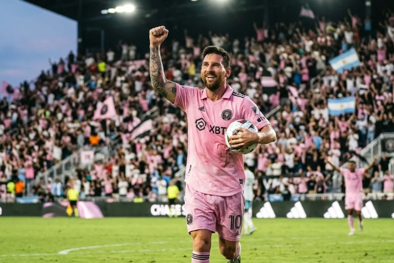 Lionel Messi smiling and celebrating a goal in his pink Inter Miami jersey, holding a soccer ball with one fist raised in front of a packed stadium crowd.