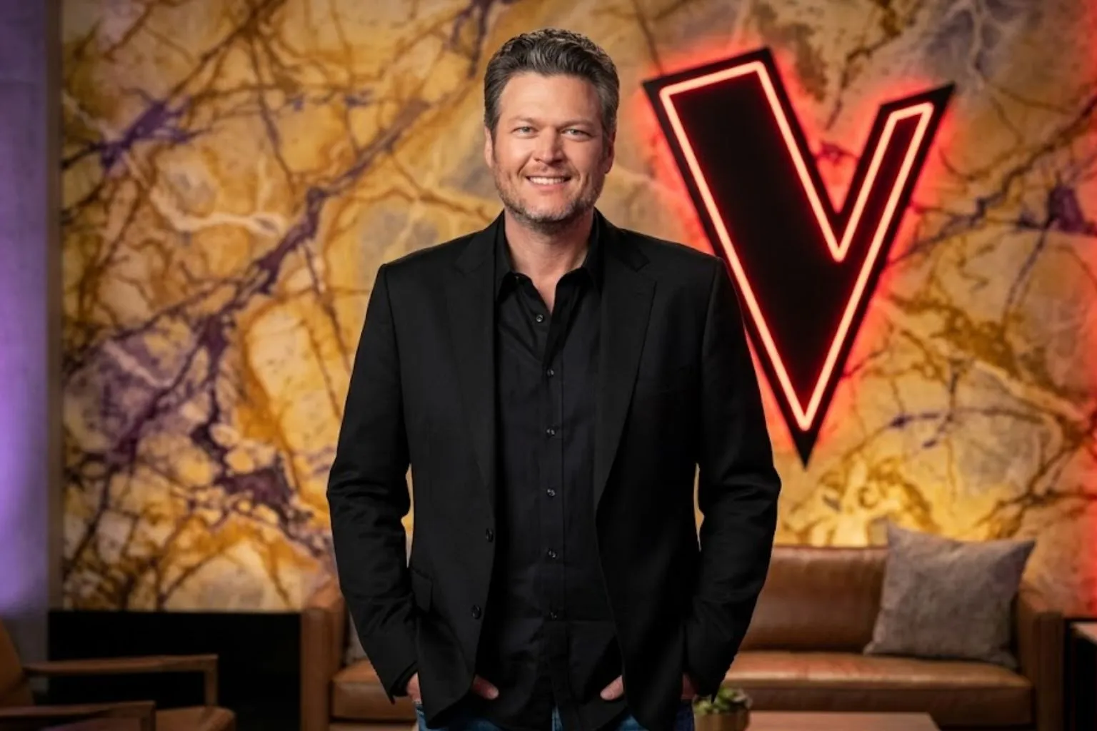 A realistic portrait of country music star Blake Shelton smiling in a black blazer and button-down shirt, standing in front of a neon 'V' sign on an entertainment studio set.