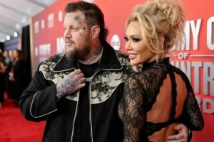 A candid, side-angle red carpet photo of musician Jelly Roll, showing his face tattoos and custom black western jacket, alongside his wife Bunnie Xo, who is wearing a black lace dress with an open back.