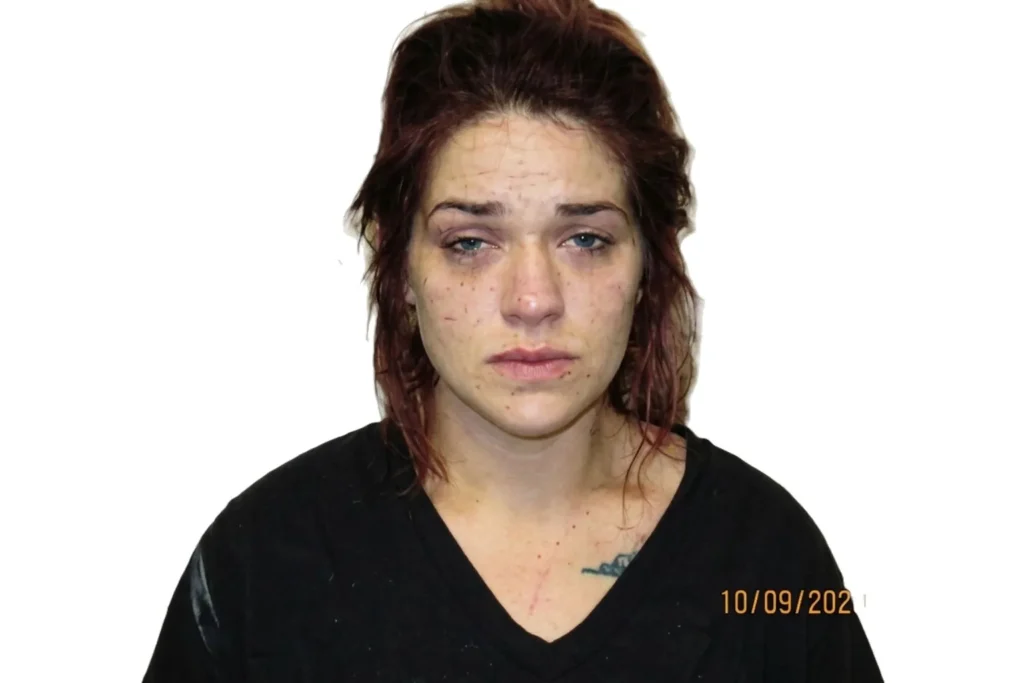 A candid booking photograph of a woman with red hair and blue eyes, showing injuries to her face, standing against a cinder block height chart.