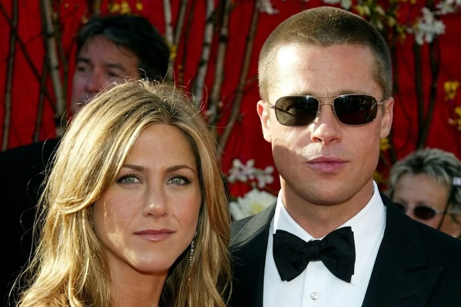 A realistic close-up red carpet portrait of Jennifer Aniston looking forward with green eyes alongside Brad Pitt wearing aviator sunglasses, a tuxedo, and a black bow tie against a vibrant red backdrop.