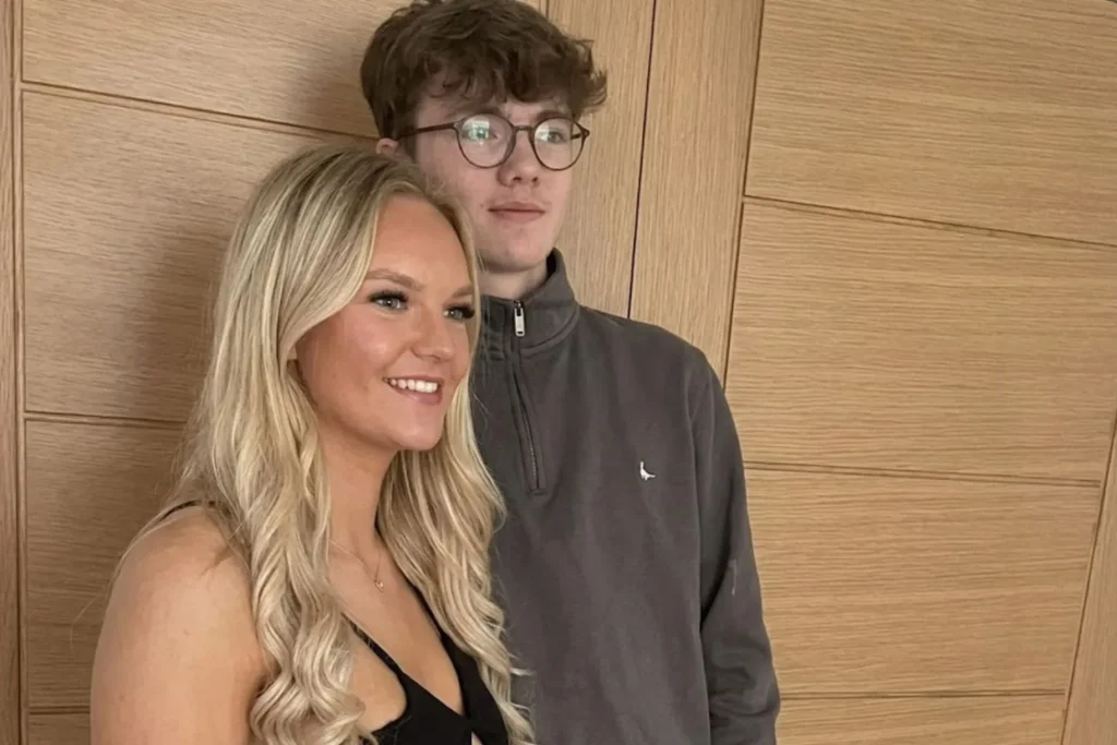 A realistic candid photograph taken from a side angle of a smiling blonde woman in a black dress standing next to a young man with brown curly hair and glasses wearing a gray pullover against a paneled wooden door.