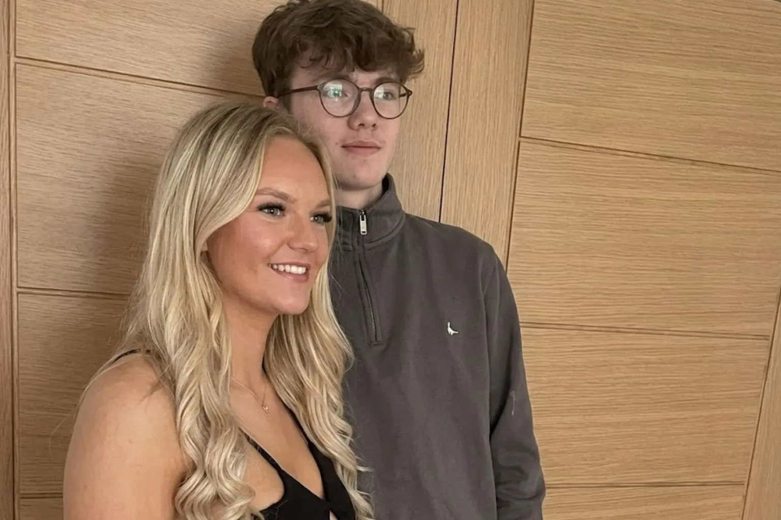 A realistic candid photograph taken from a side angle of a smiling blonde woman in a black dress standing next to a young man with brown curly hair and glasses wearing a gray pullover against a paneled wooden door.