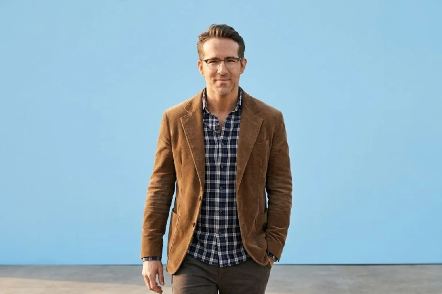 Ryan Reynolds walking outdoors in a brown corduroy blazer and plaid shirt against a clear sky blue wall.