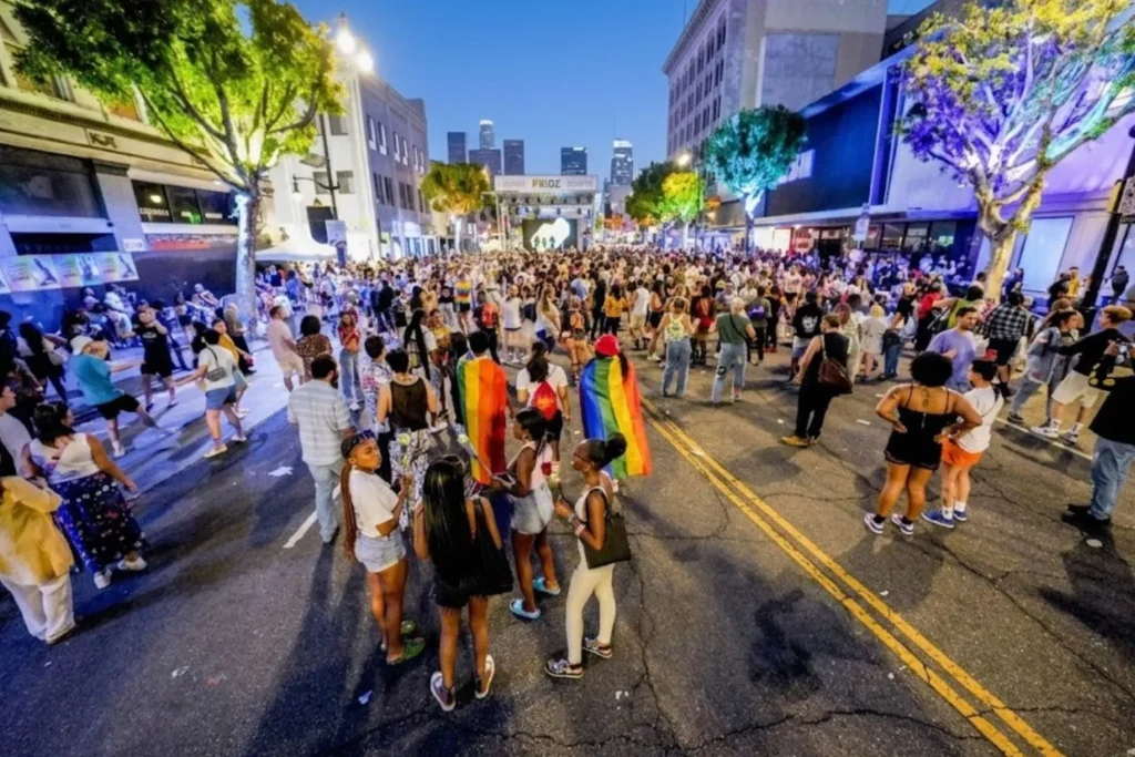 A large crowd gathered on a city street at dusk for a Pride event, with people wearing rainbow flags and a lit-up stage framed by skyscrapers in the distant background.