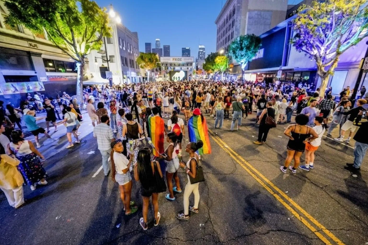 A large crowd gathered on a city street at dusk for a Pride event, with people wearing rainbow flags and a lit-up stage framed by skyscrapers in the distant background.