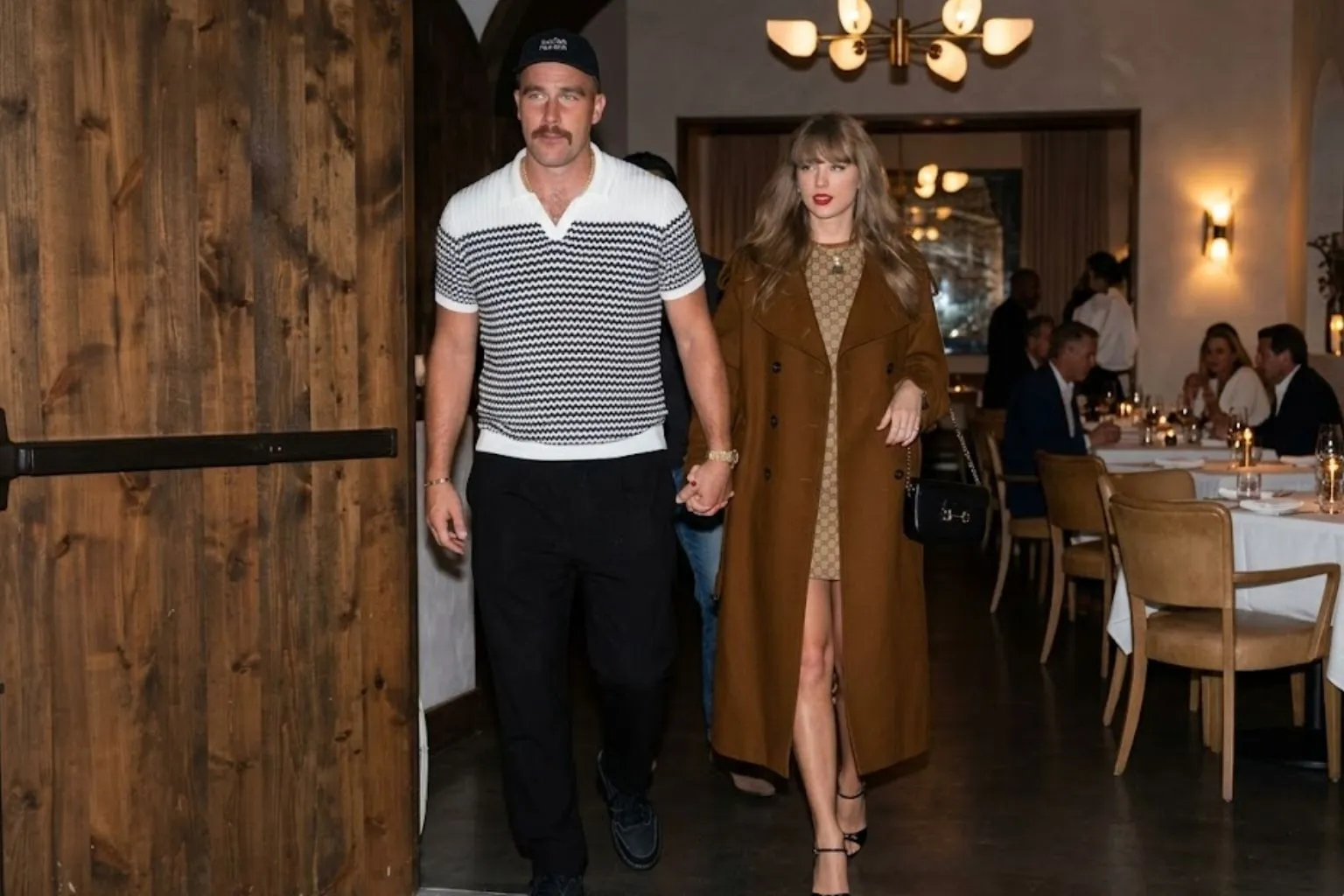 Travis Kelce and Taylor Swift holding hands while walking out of a dimly lit restaurant.