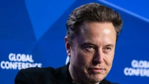 Tech billionaire Elon Musk gesturing dynamically on stage next to an entertainment display showcasing controversial streaming trends.