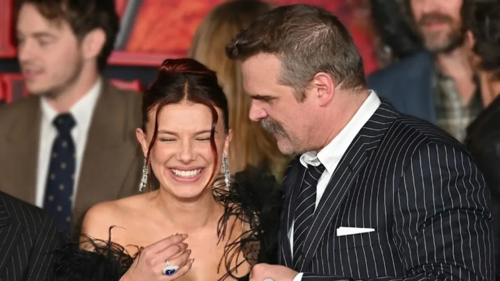 Millie Bobby Brown and David Harbour posing together at a high-profile Hollywood red carpet event, smiling for cameras.