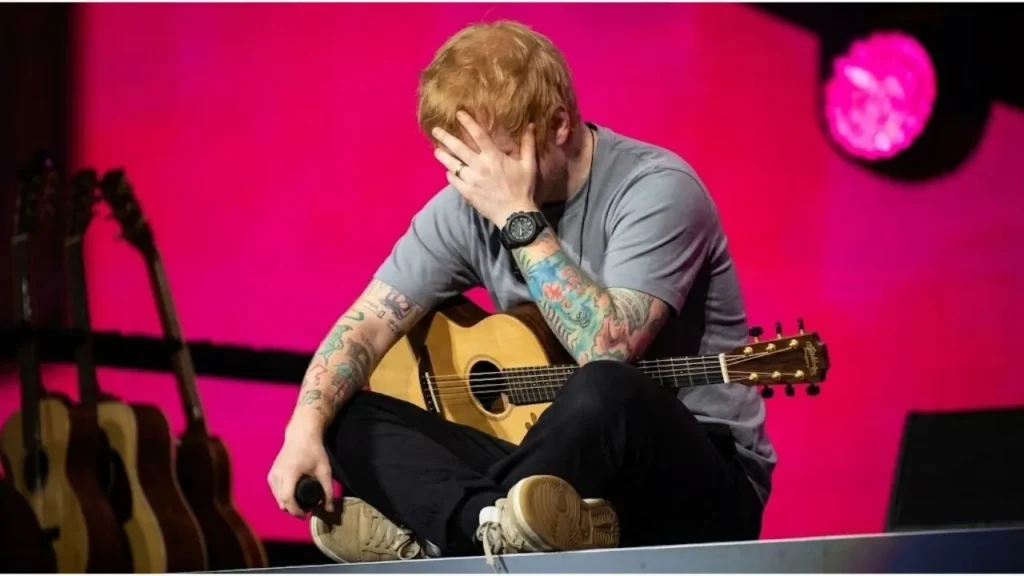 Ed Sheeran performing solo on stage with an acoustic guitar under bright stadium lights